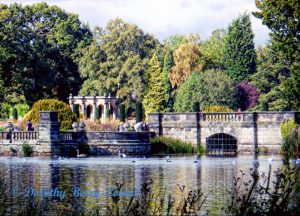 A peaceful scene featuring a lake, a stone bridge and a viewing area where people can stand and watch the wildfowl on the water. All against a tree-lined background