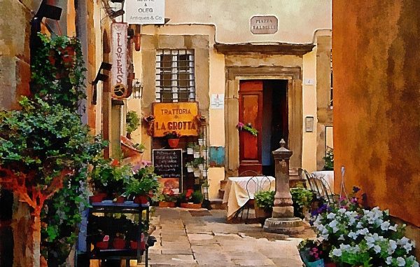 A look down an alleyway past planted containers full of flowers to a courtyard beyond. A table and chairs sits next to a restaurant sign and there is a water fountain. A half open door is just behind the table. The alleway goes from shade into the light highlighting the traditional Italian architecture in the background.