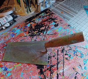 A plasterers tool, a finishing trowel with a wooden handle sits on an abstract painting of predominantly red, turquoise and black. The painting sitson a table with a patterned table cloth and in the background can been some paint supplies.